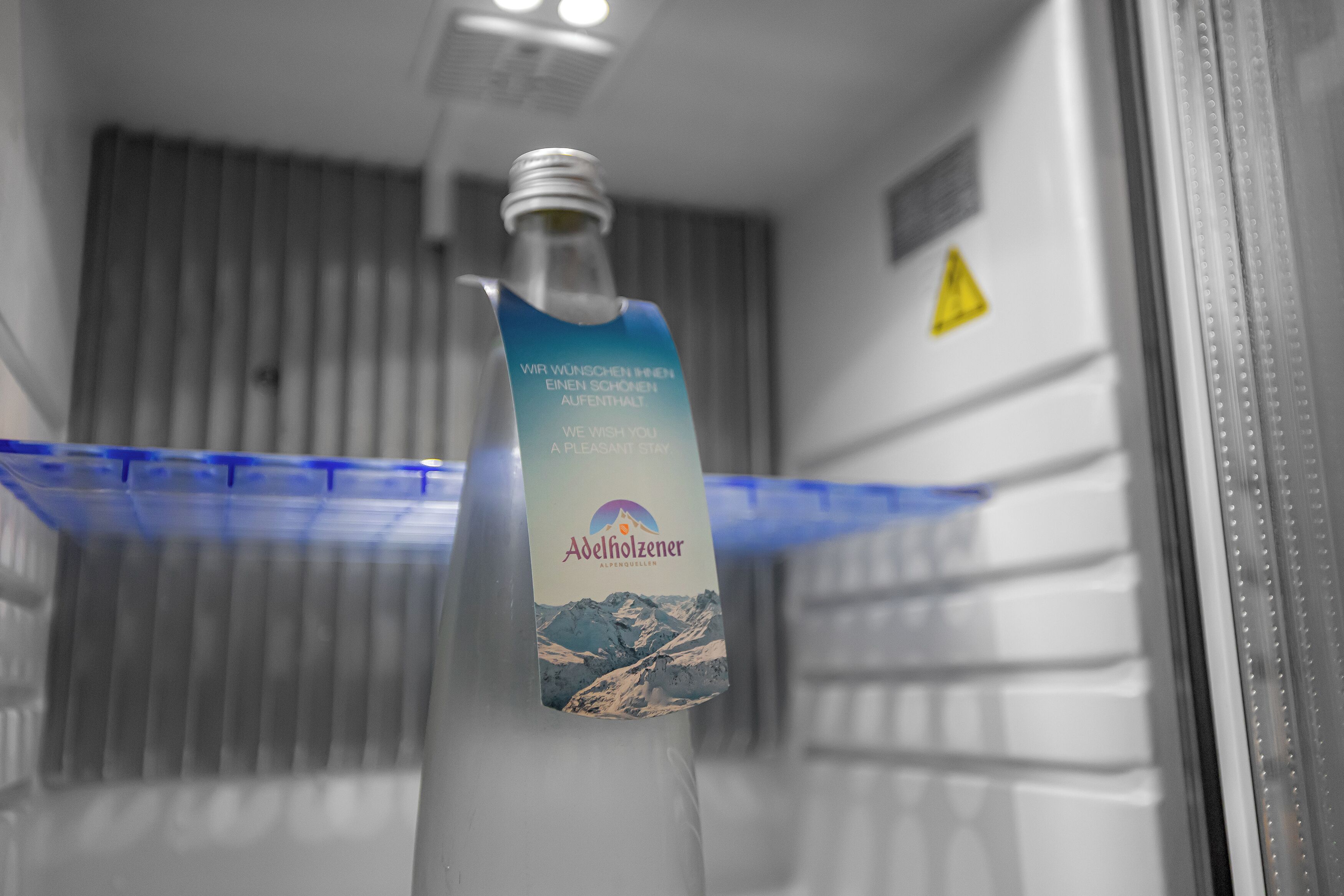 A bottle of Adelholzer beer in a refrigerator with a blue interior and a warning sign on the door.