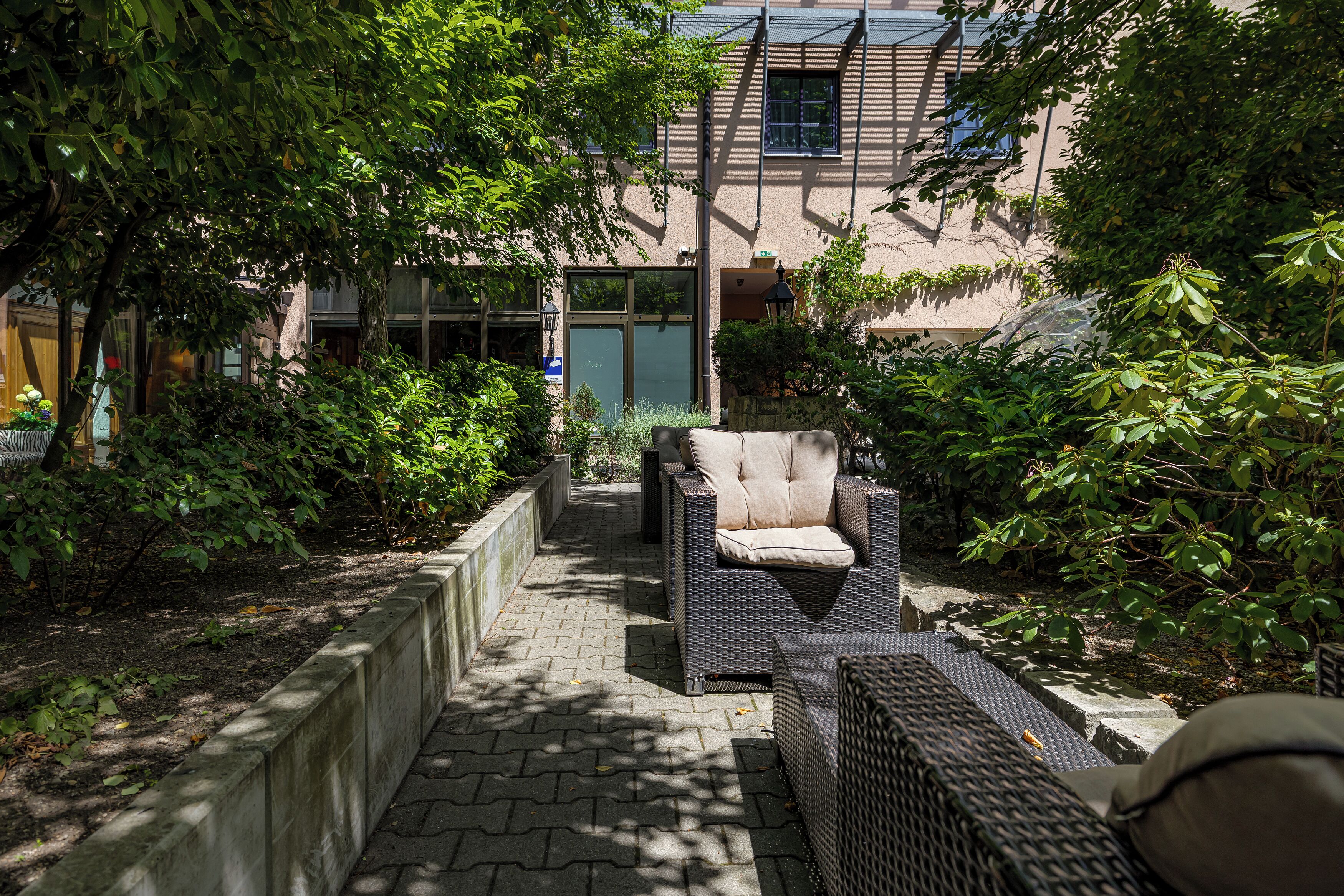 A pathway leading to a seating area with a chair and a sofa, surrounded by greenery and a building with large windows in the background.