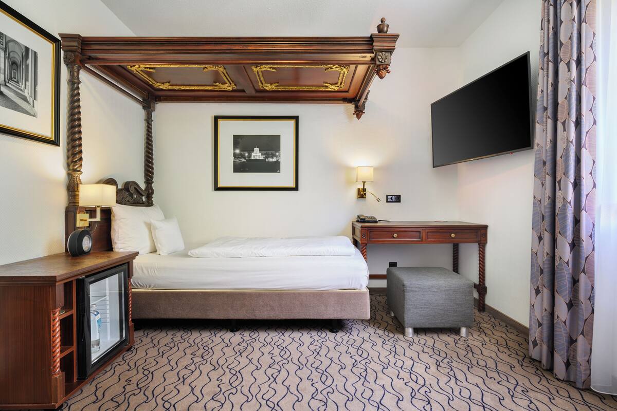 Habitación de hotel con cama, escritorio, televisor, un cuadro enmarcado, lámpara y alfombra estampada.
