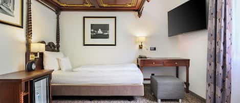 Habitación de hotel con cama, escritorio, televisor, un cuadro enmarcado, lámpara y alfombra estampada.