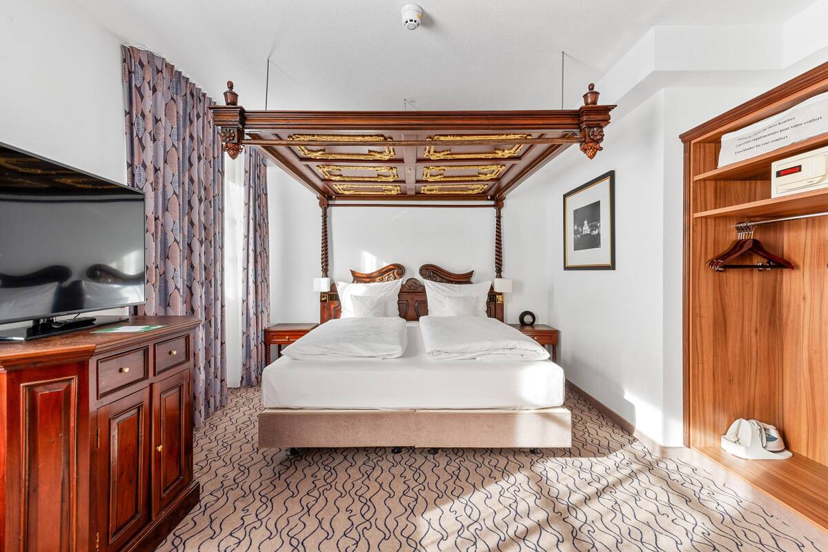 Un dormitorio con una cama con dosel, un televisor sobre un mueble de madera, cortinas estampadas y un cuadro en la pared.