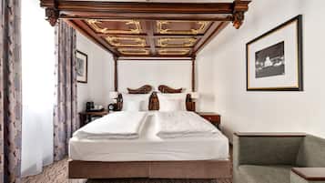 A large bed with a wooden headboard and a canopy, a nightstand with a lamp, a framed picture on the wall, and a green armchair.