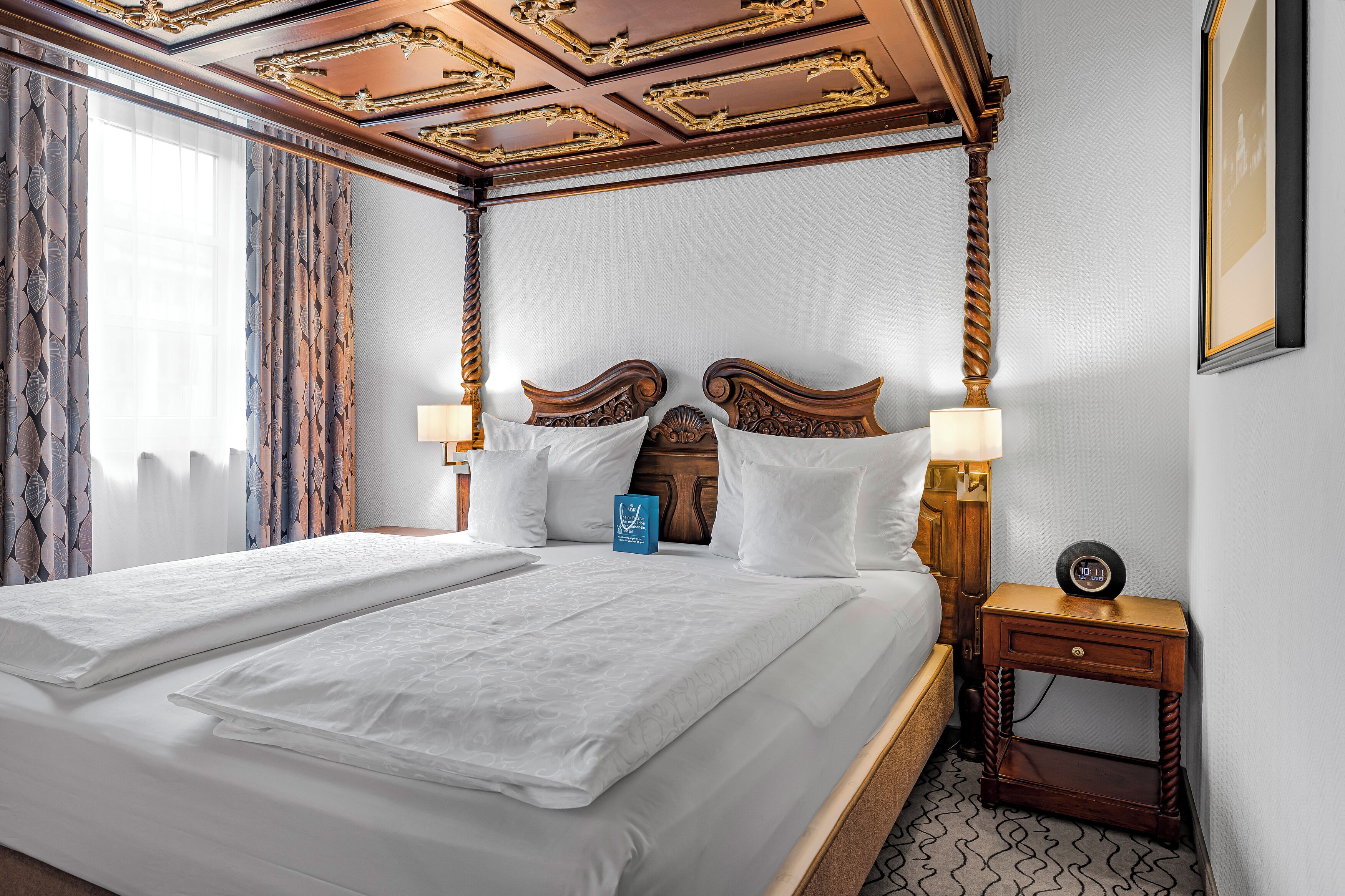 Habitación de hotel con una cama grande, dos lámparas de noche, un cabecero de madera, una mesita de noche y un reloj.
