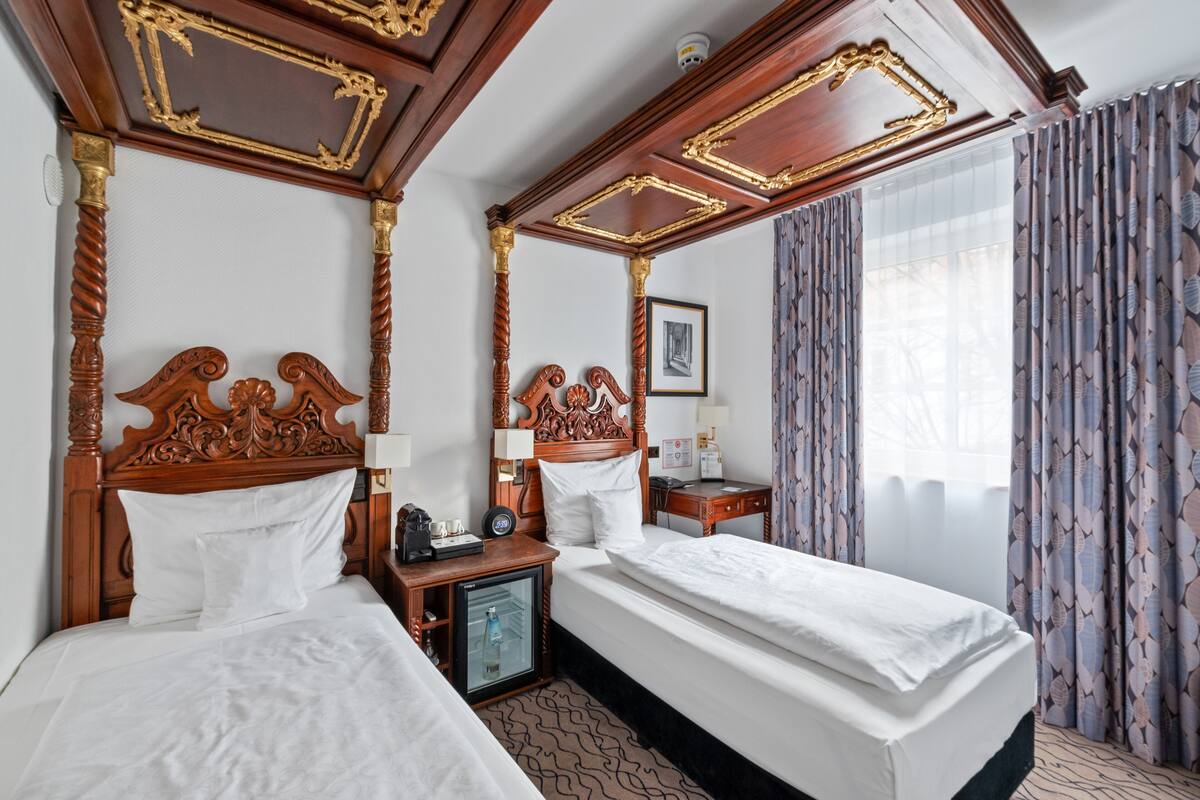 Una habitación de hotel con una cama grande, una cama más pequeña, una mesita de noche, una lámpara, un cuadro enmarcado y cortinas.