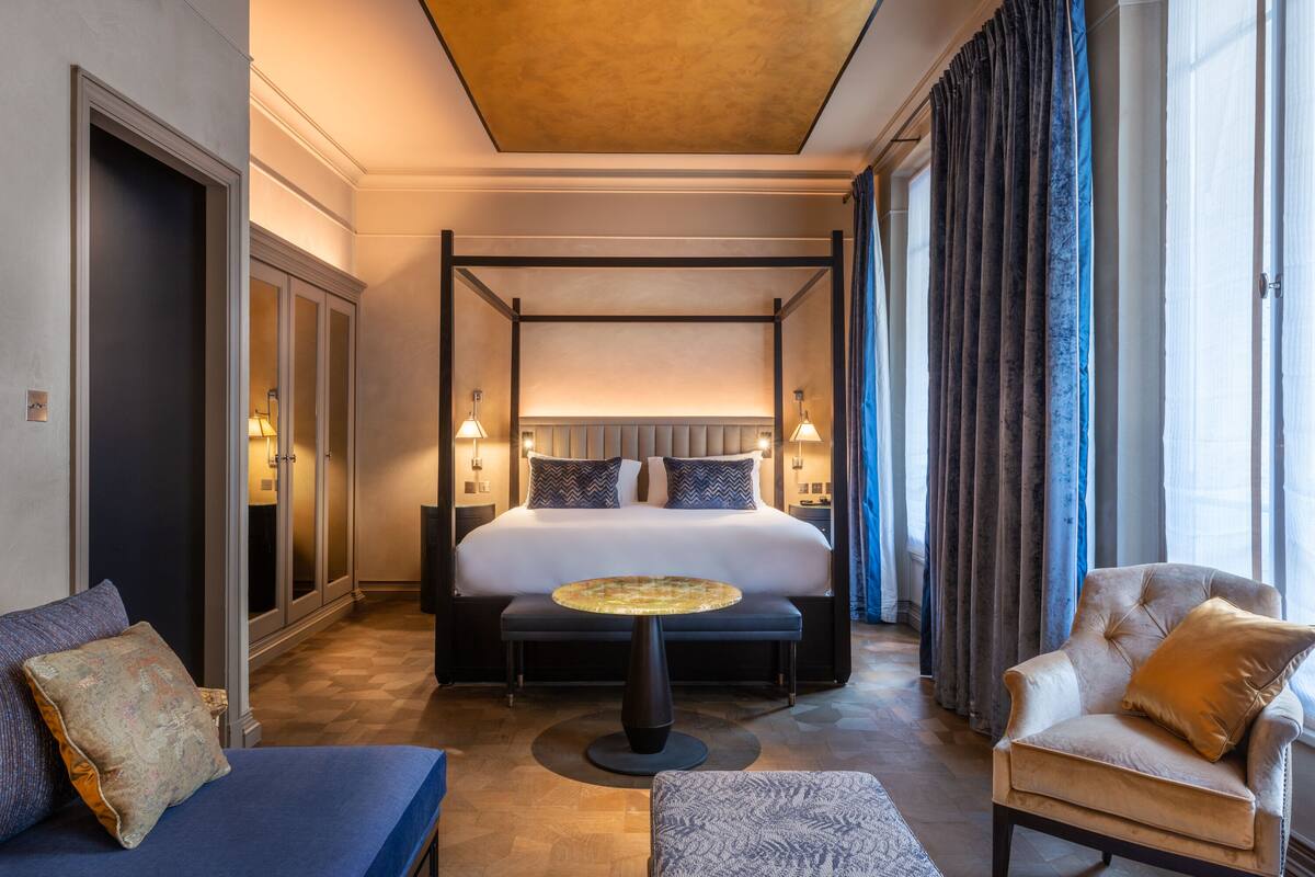 A spacious hotel room with a four-poster bed, a round coffee table, a sofa, and a chair.