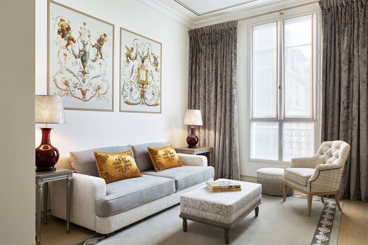 A living room with a grey sofa, two gold pillows, a patterned ottoman, a red lamp, and a chair.