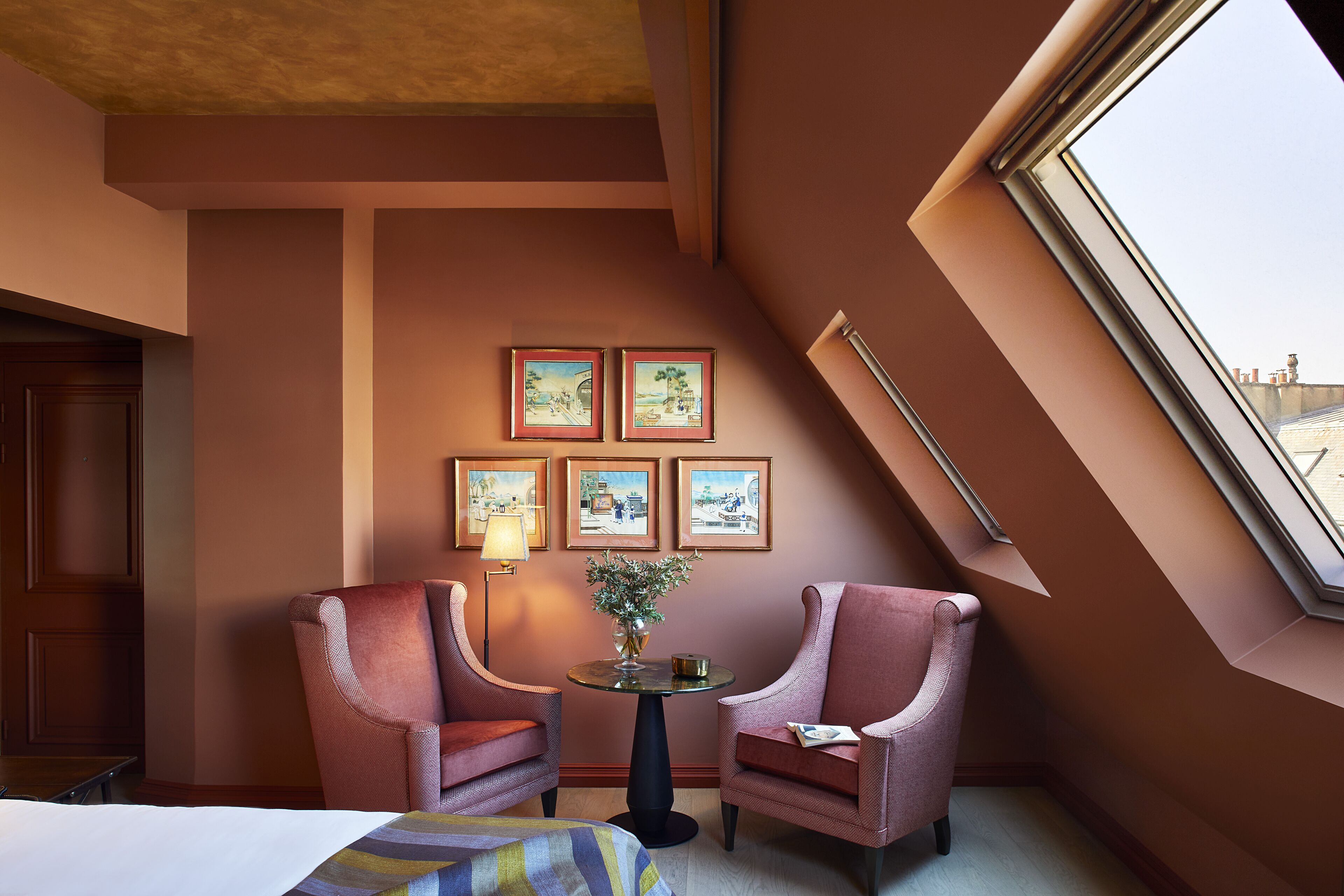 Habitación de hotel con techo inclinado, dos sillones, una mesita redonda con un florero y tres cuadros en la pared.