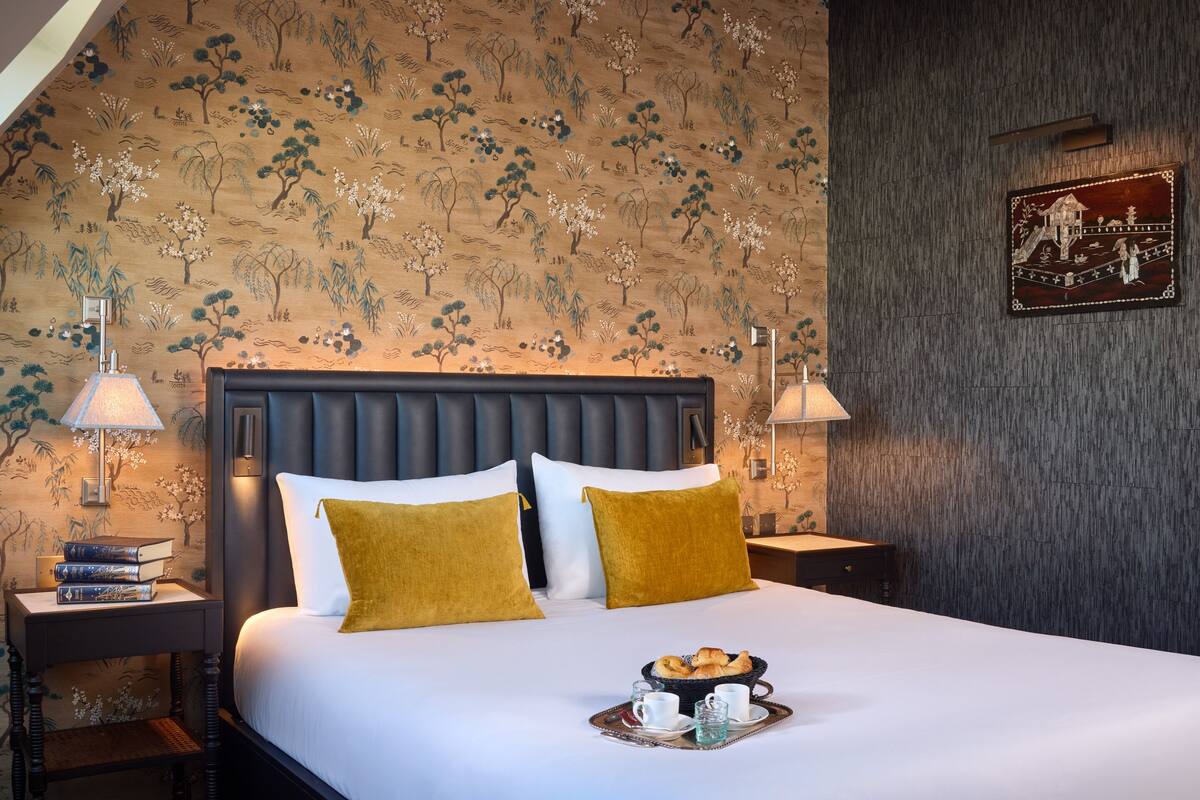 A hotel room with a large bed, two bedside tables, a nightstand, a wall with a floral pattern, and a framed picture.