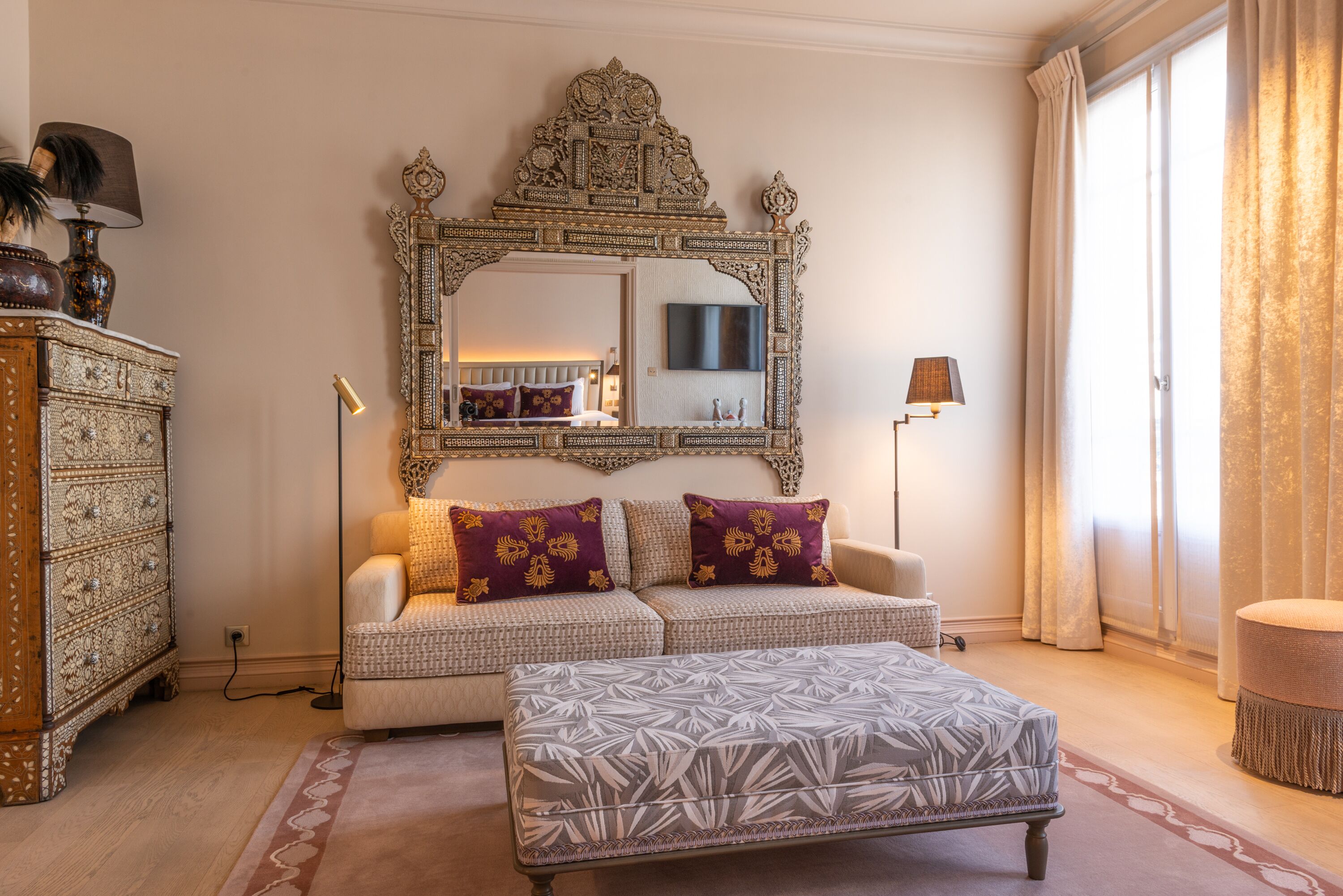 A living room with a patterned sofa, a matching ottoman, a decorative mirror, and a traditional wooden dresser.