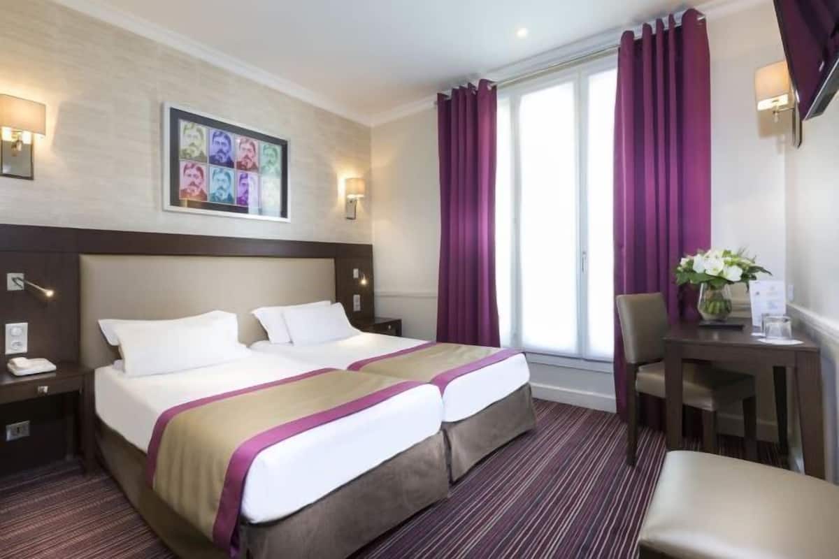 A hotel room with a double bed, a desk, a chair, a window with purple curtains, and a wall-mounted picture.