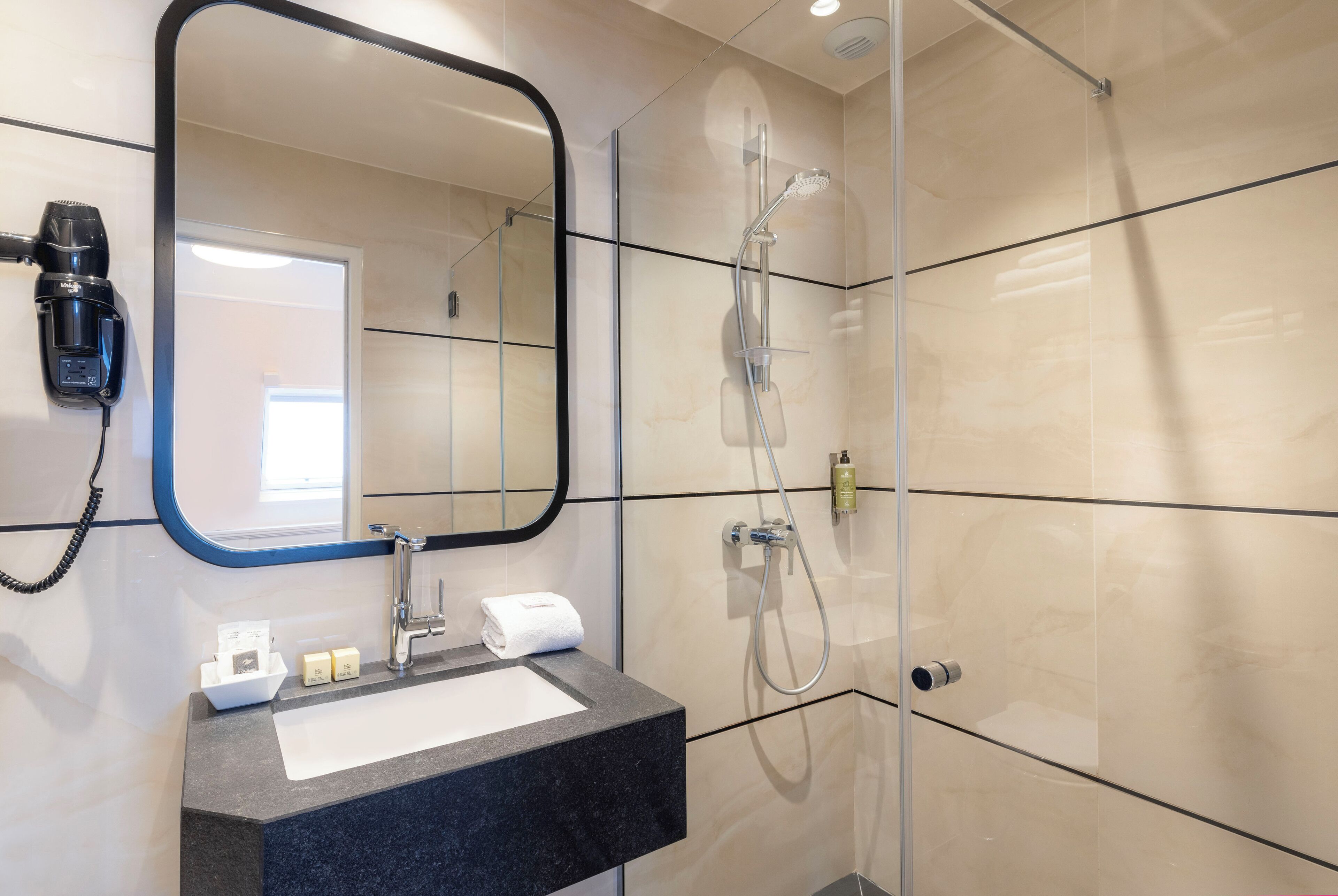 Une salle de bain moderne dotée d’une douche à parois de verre, d’un lavabo à poser noir, d’un miroir et d’un sèche-cheveux fixé au mur.