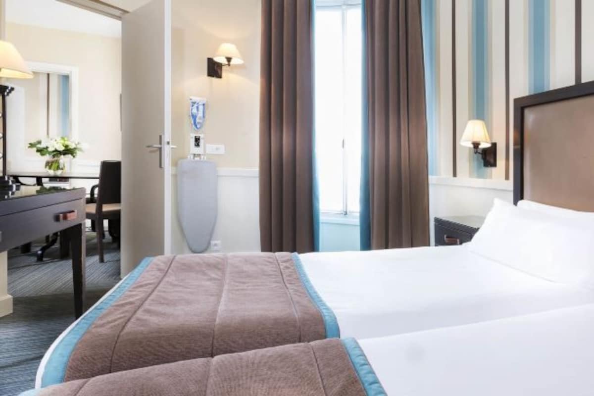 A hotel room with a bed, a desk, a chair, a lamp, and a window with curtains.