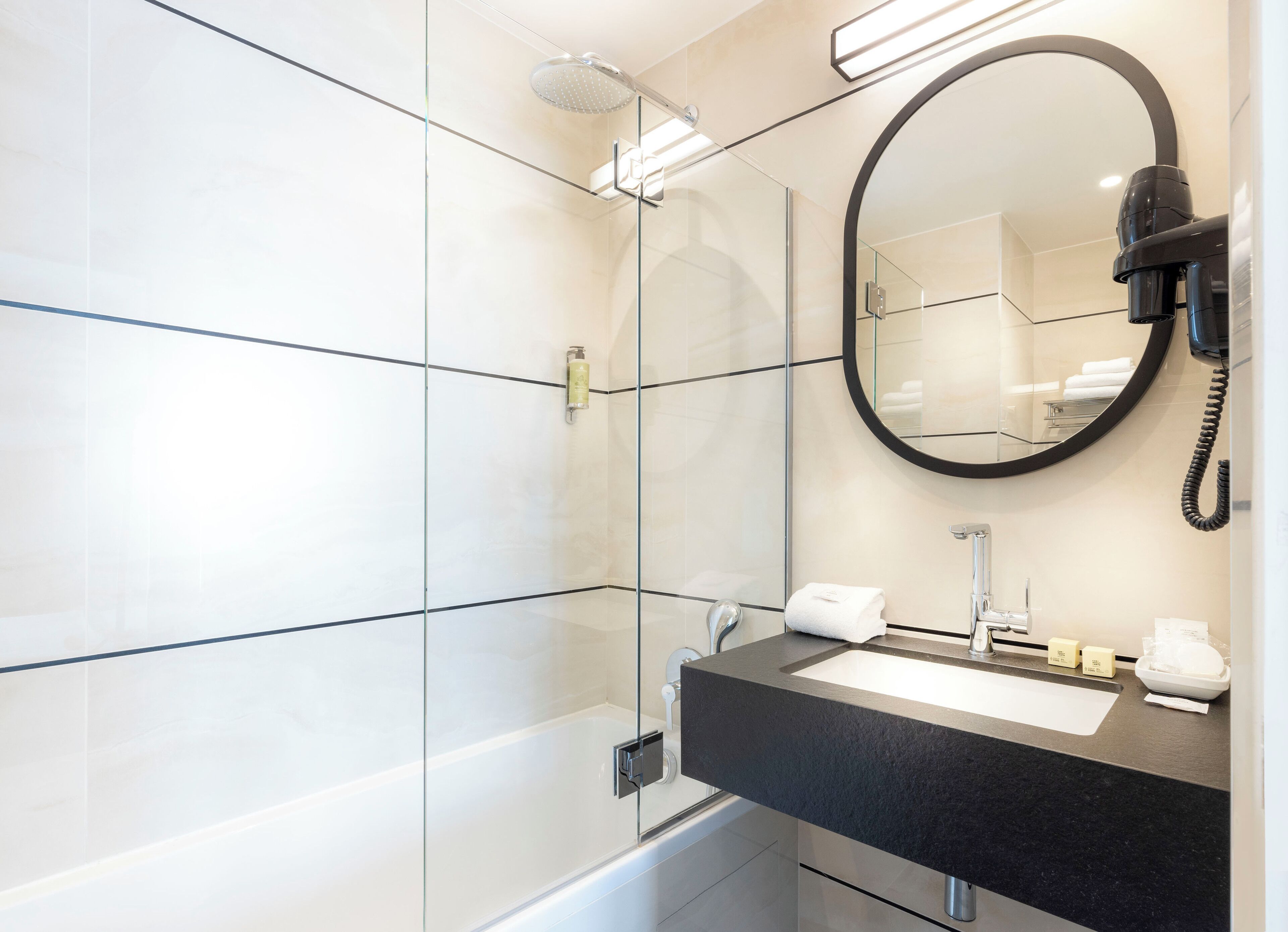 A modern bathroom with a glass shower enclosure, a black countertop sink, a round mirror, and a hairdryer mounted on the wall.
