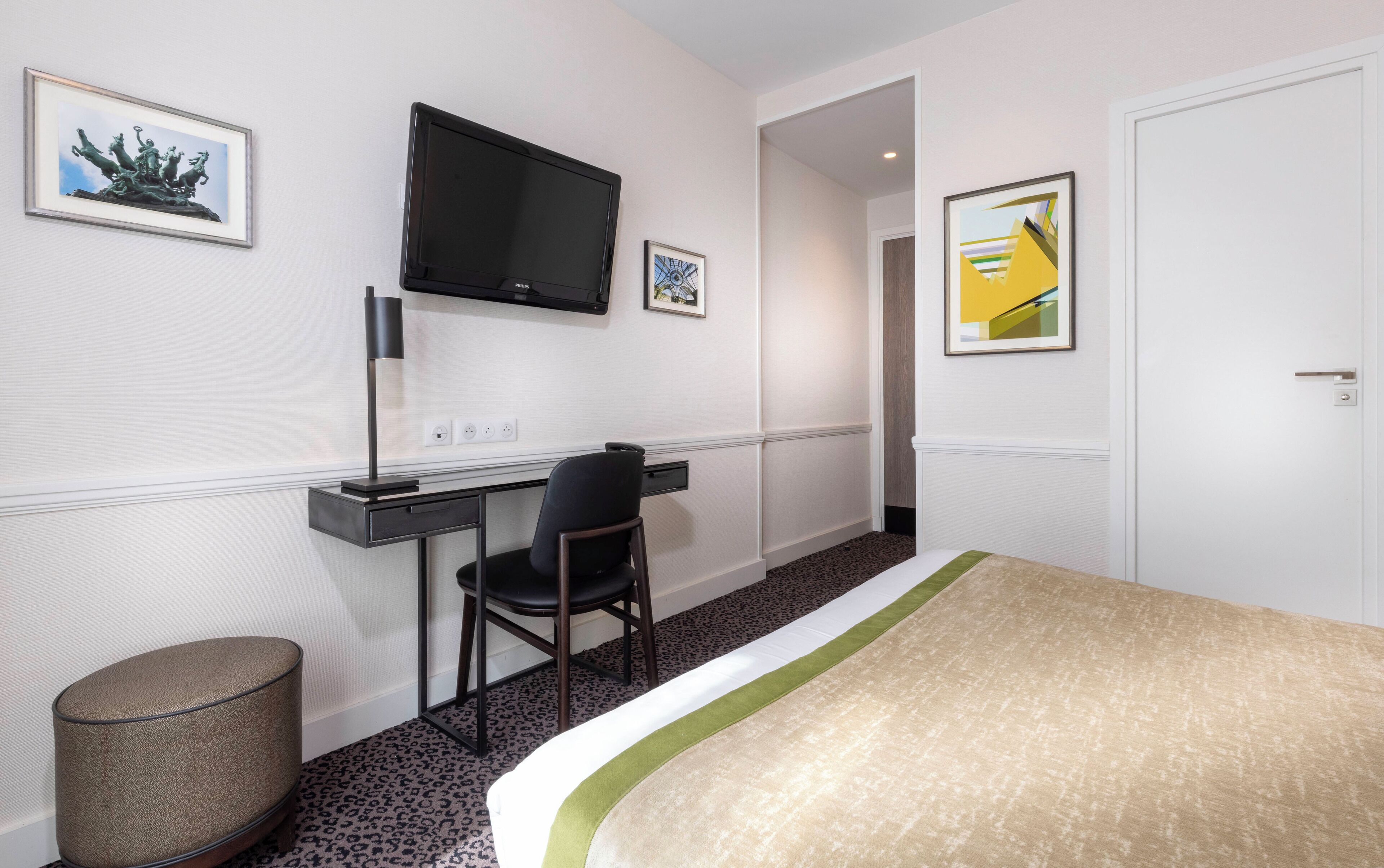 A hotel room with a bed, a desk with a chair, a television mounted on the wall, and two framed pictures.