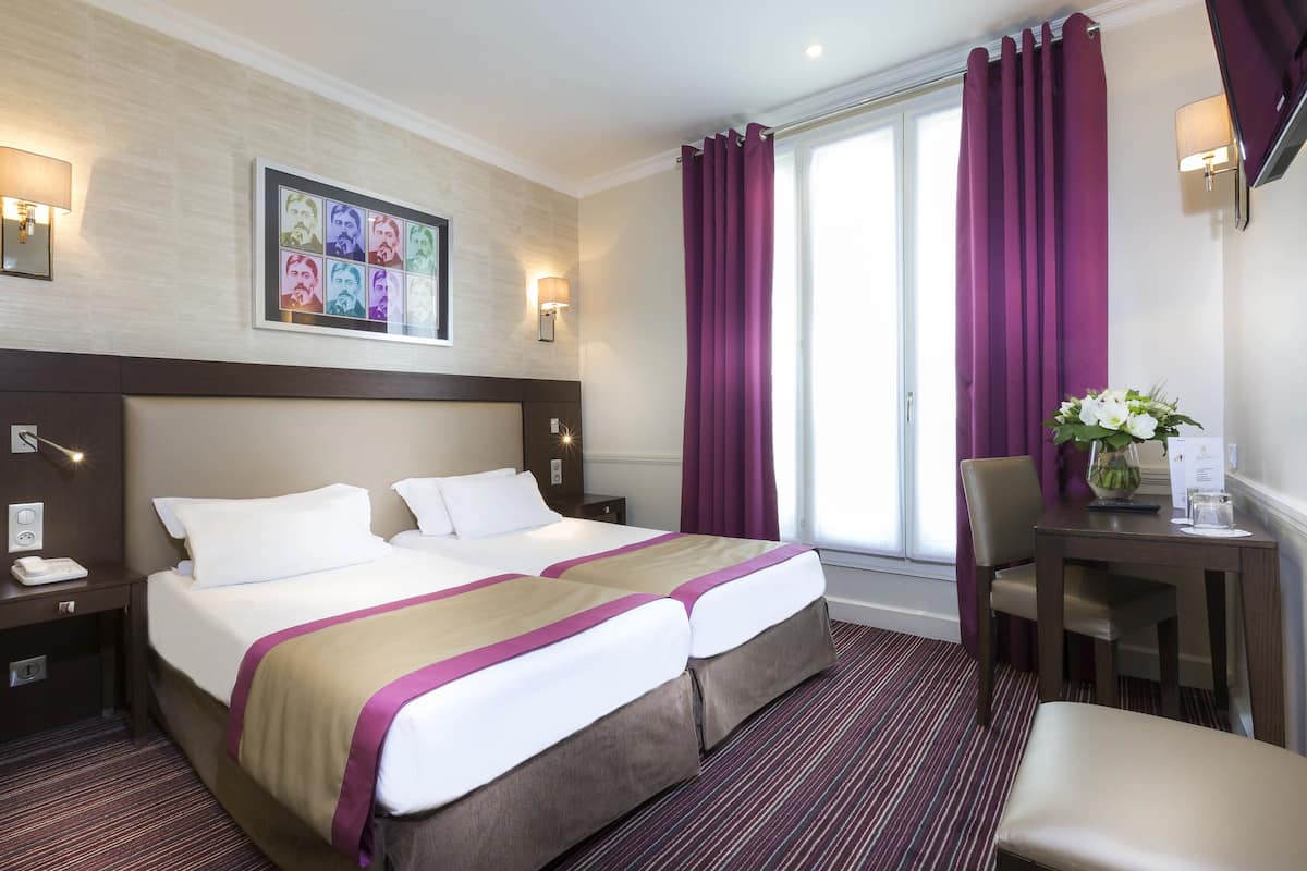 A hotel room with a large bed, two bedside tables, a desk with a chair, a window with purple curtains, and a wall-mounted artwork.