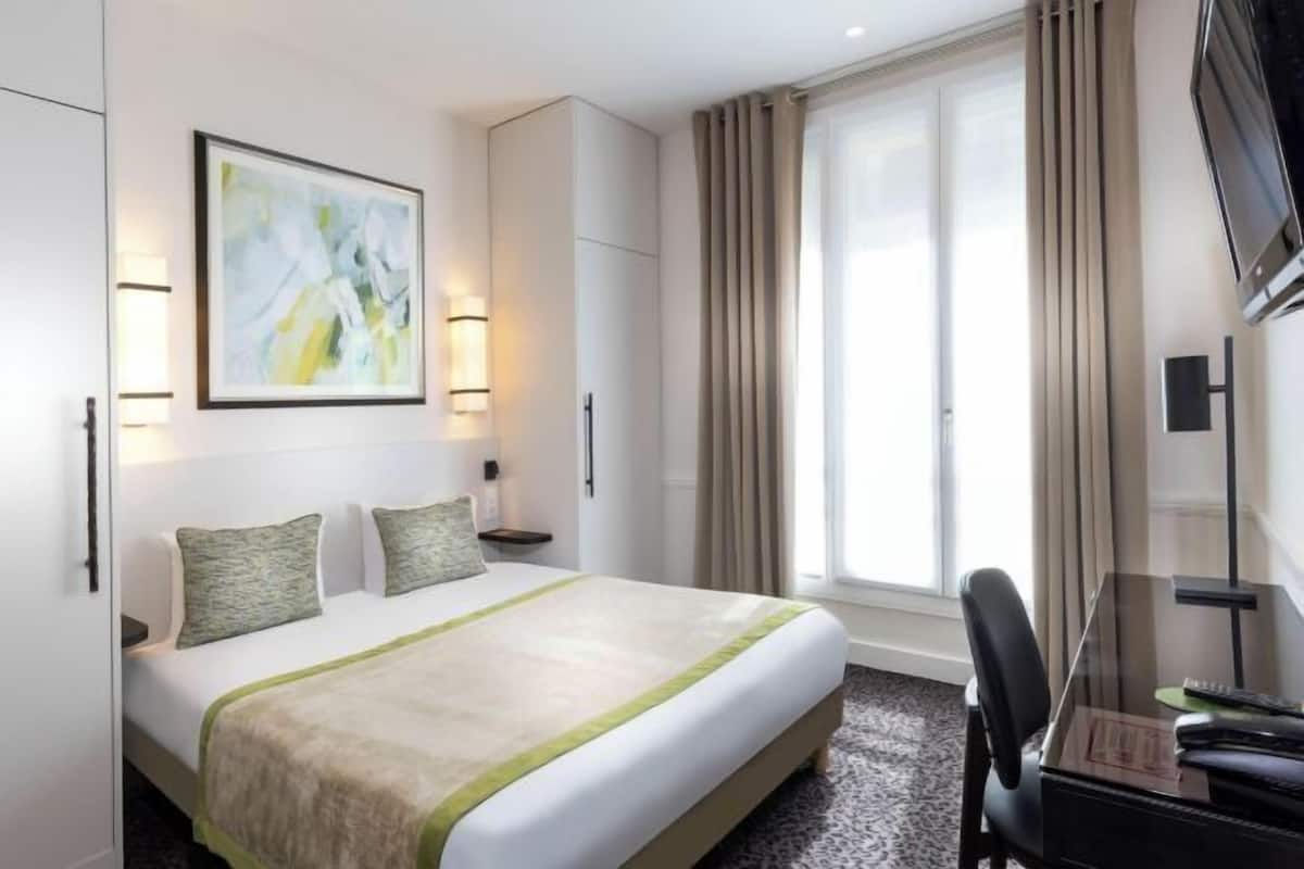 A hotel room with a bed, a desk, a chair, a TV, and a window with curtains.