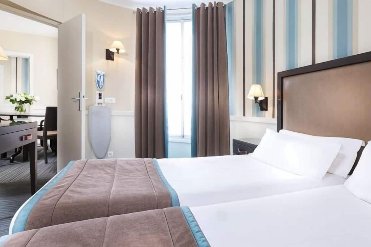 A hotel room with two beds, a desk, a chair, a lamp, and a window with curtains.