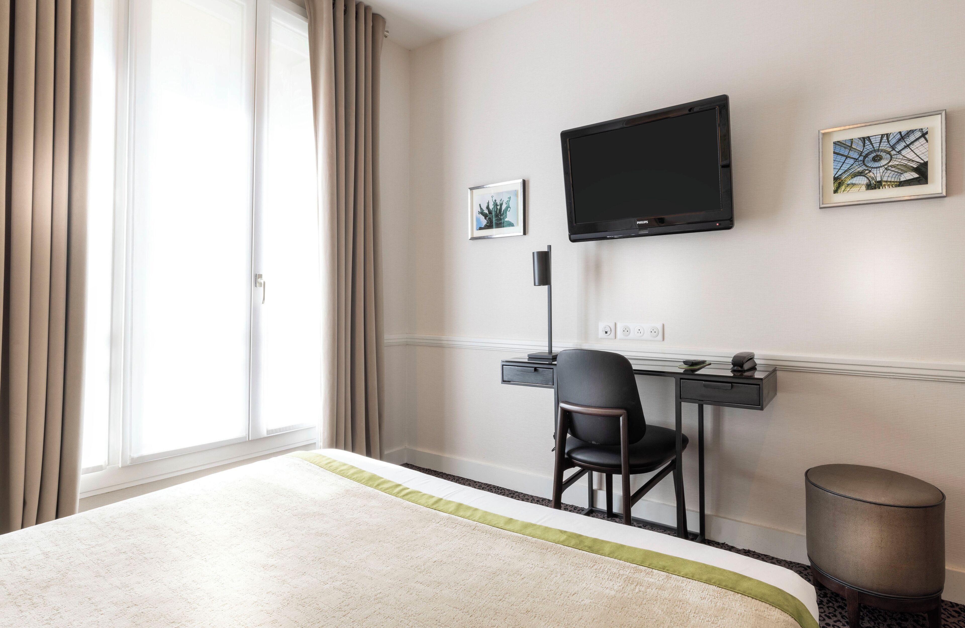Quarto de hotel com cama, escrivaninha com cadeira, televisão e duas imagens emolduradas na parede.