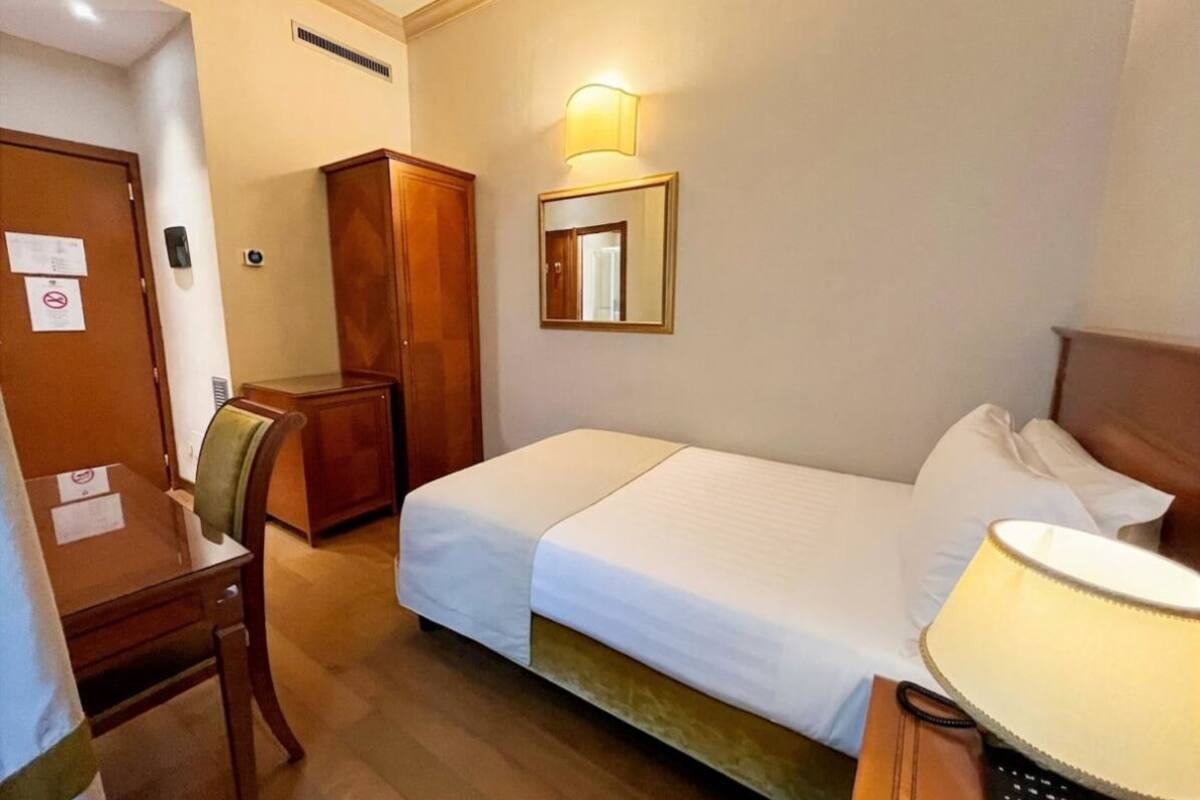A hotel room with a bed, a desk, a chair, a wardrobe, a mirror, and a lamp.