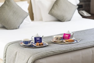 Colazione ordinata su un letto con testiera grigia imbottita, che include teiera, tazze e pasticcini.