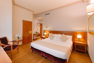 Camera d'albergo con un letto grande, una testiera in legno, due comodini con lampade, una sedia di legno e una scrivania con un vaso di fiori.