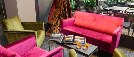 Un área de descanso moderna con un sofá rojo, sillones verdes y rosados, y una mesa de centro con una revista y bebidas.