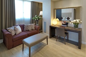 Een hotelkamer met een bruine bank, een witte bankje, een bureau met stoel, een grote spiegel en een vaas met bloemen.