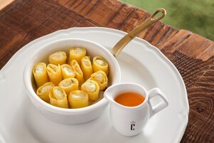 Een witte schaal met rolletjes gebak, met een gouden lepel op een wit bord, vergezeld van een klein wit kopje saus.