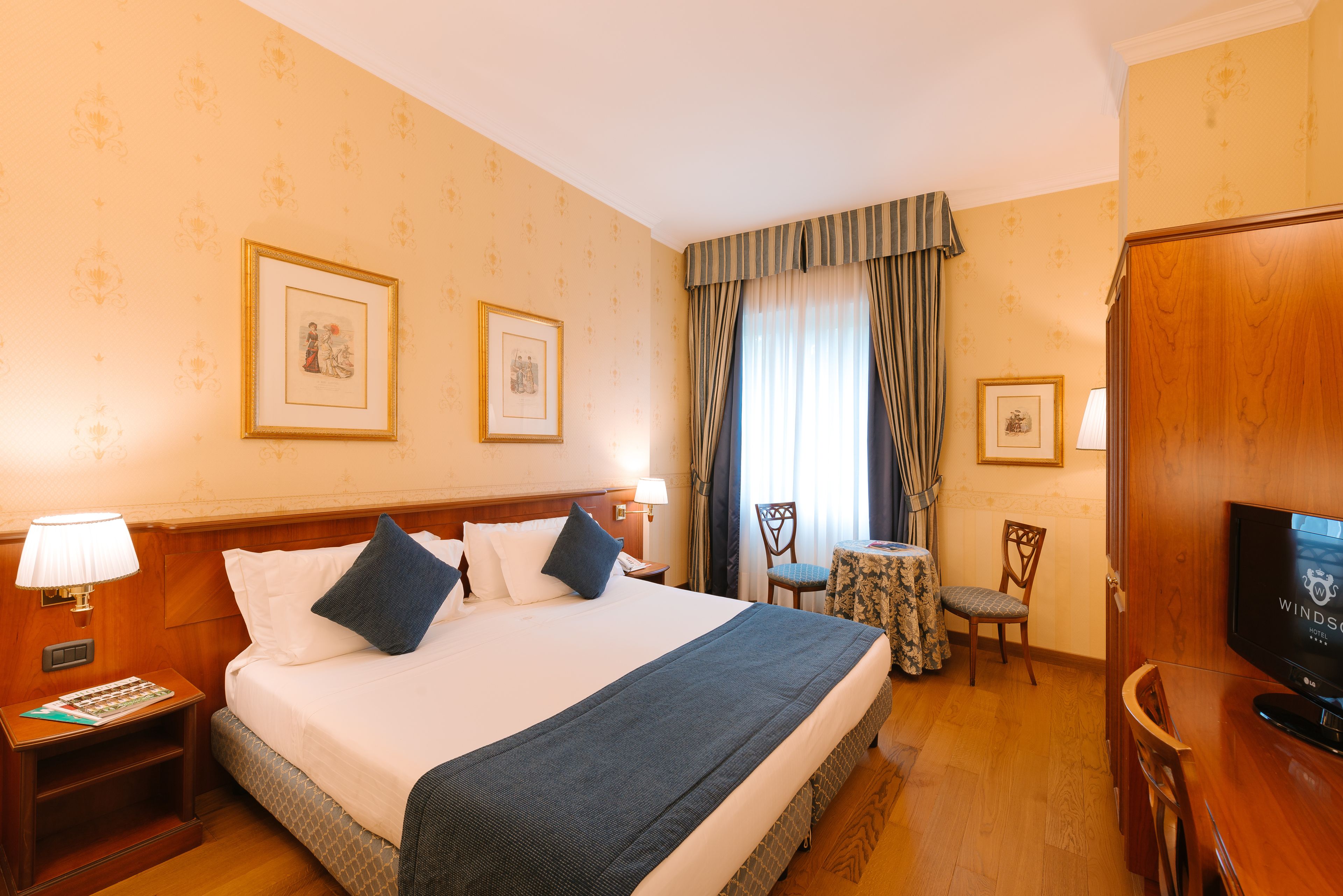 Ein Hotelzimmer mit einem großen Bett, einem Fernseher, einem kleinen Tisch mit einem Stuhl und gerahmten Bildern an der Wand.