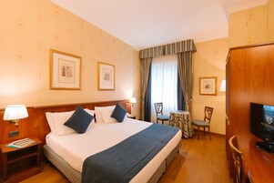 Ein Hotelzimmer mit einem großen Bett, einem Fernseher, einem kleinen Tisch mit einem Stuhl und gerahmten Bildern an der Wand.