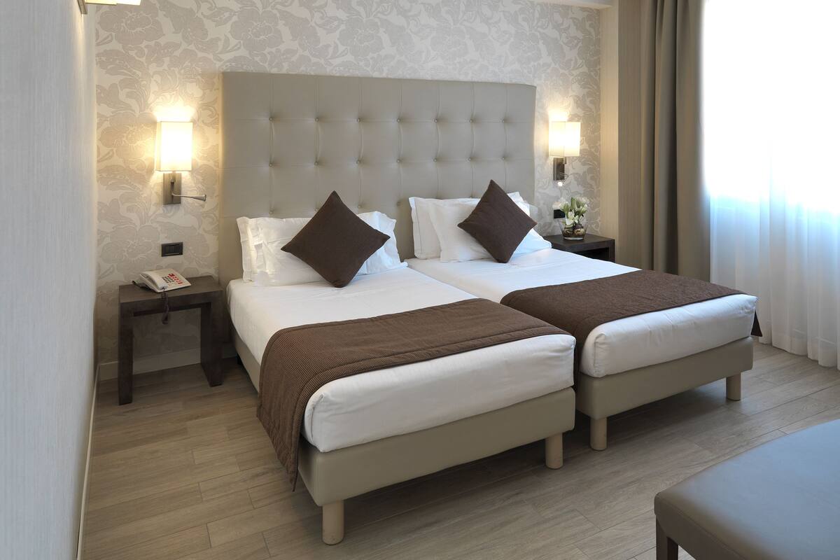 A hotel room with two beds, a large tufted headboard, bedside lamps, a small table with a phone, and a window with curtains.