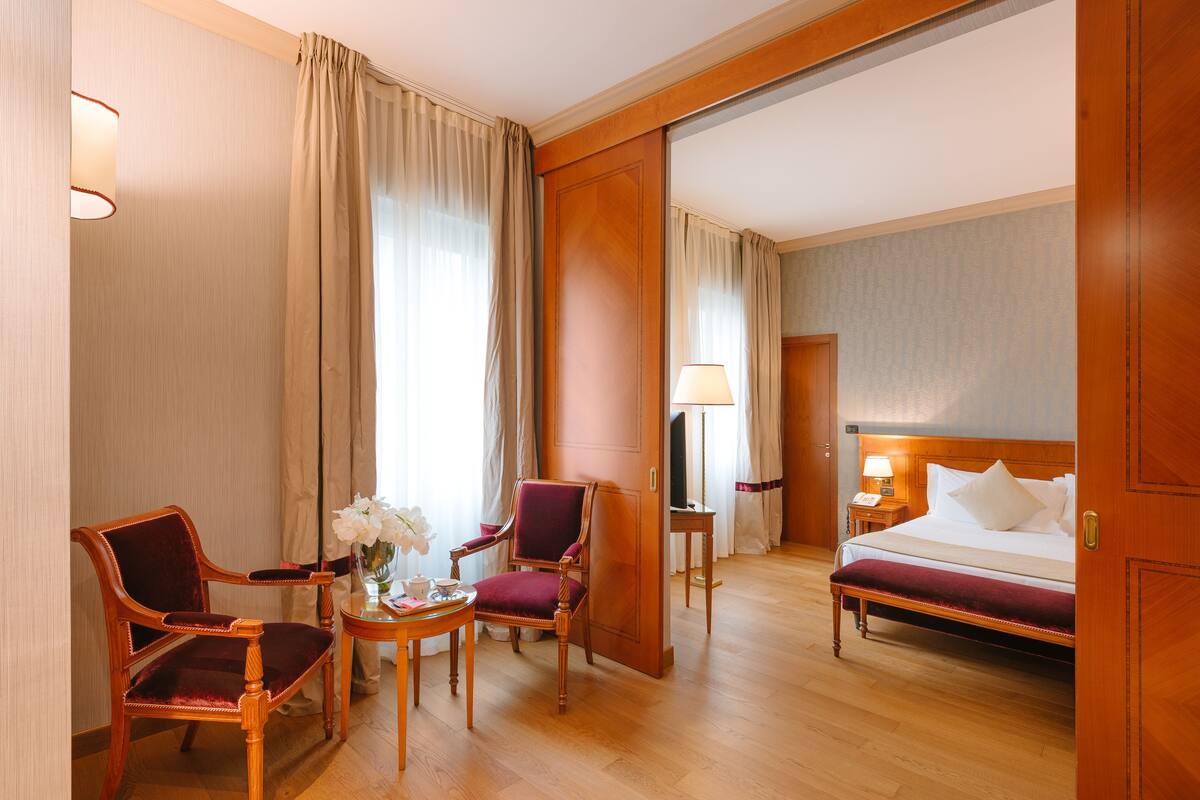 A hotel room with a bed, two chairs, a small table with a vase of flowers, a nightstand with a lamp, and a wooden door.