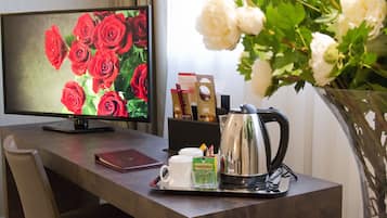 Un escritorio con un monitor de computadora mostrando rosas rojas, una cafetera, una bandeja con una taza y sobres de té, y un florero con flores blancas.