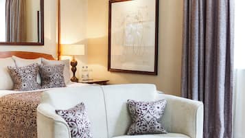 A hotel room with a bed, a white sofa, patterned pillows, a framed artwork on the wall, a bedside table with a lamp, and a window with curtains.