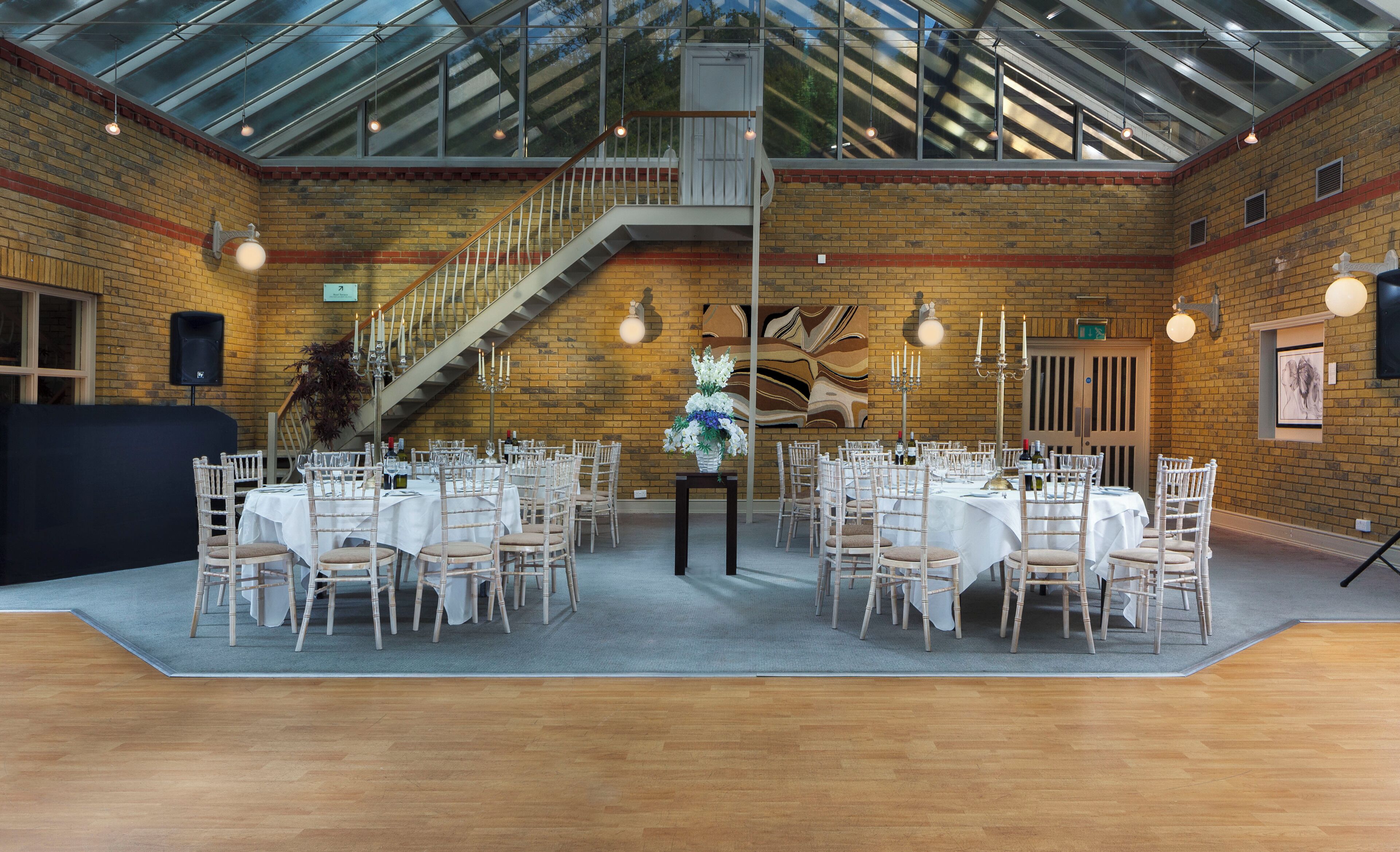 A spacious hall with a high ceiling, brick walls, and a staircase. Tables are set with white tablecloths and chairs, and there are candles and a floral centerpiece on the tables.