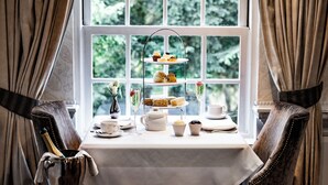 Un présentoir à trois niveaux, garni de desserts, est posé sur une table près d’une fenêtre ornée de rideaux. On y voit également un vase de fleurs et un seau à champagne.