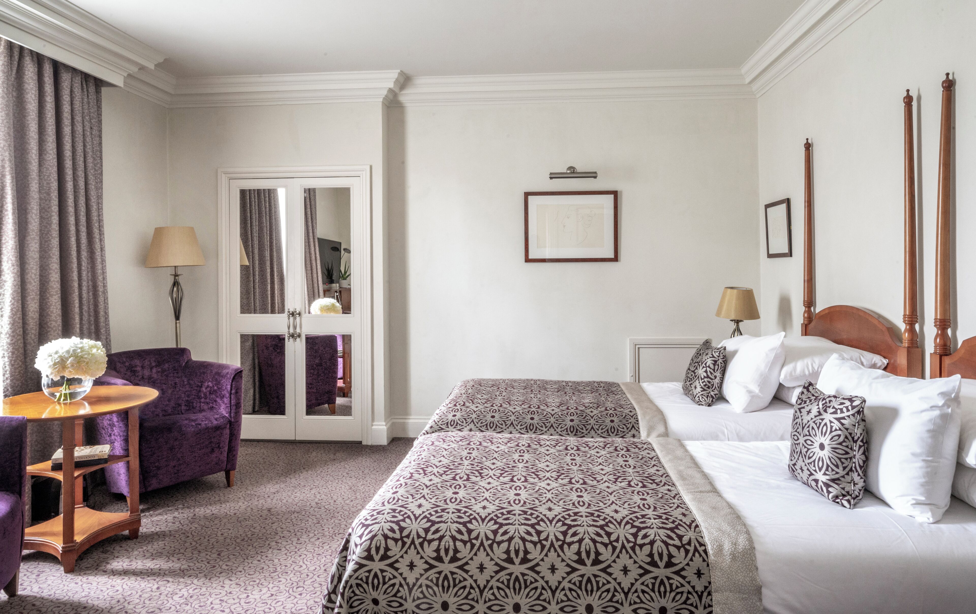 A hotel room with two beds, a sitting area with a purple chair, a small table, and a mirror.