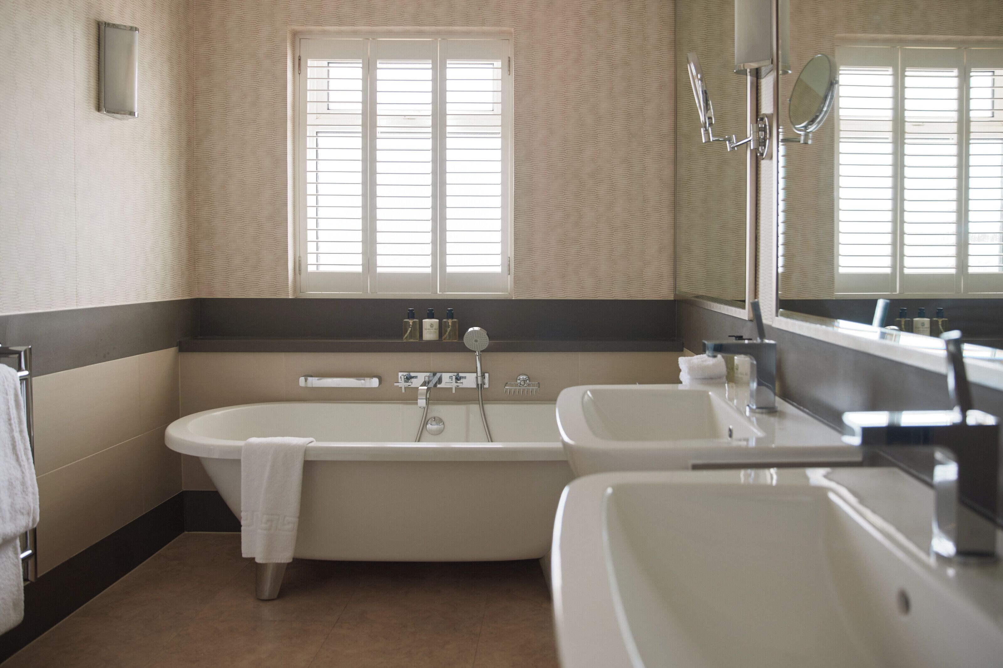 Un baño con bañera, dos lavamanos, una ventana con persianas y un espejo.