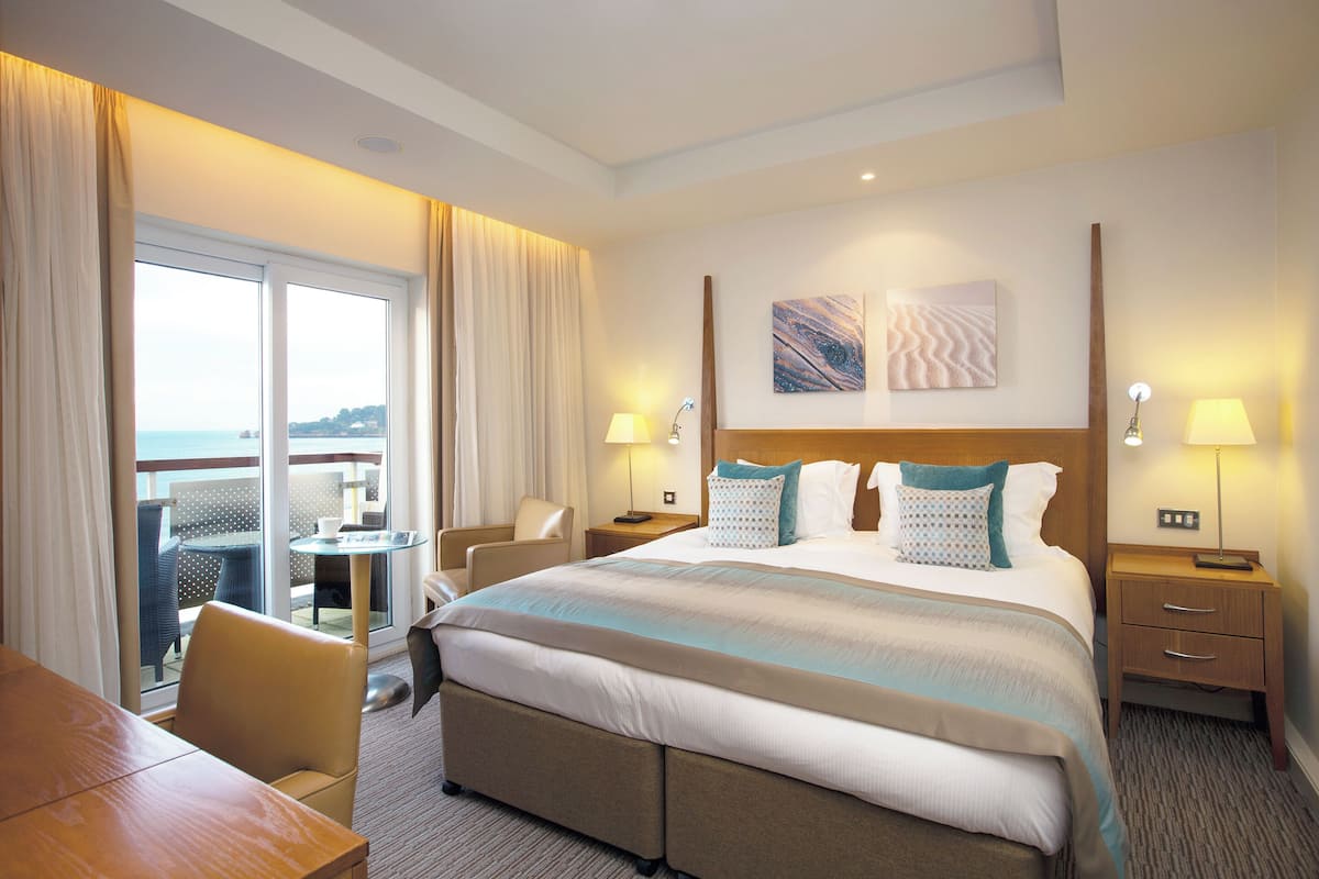 A hotel room with a large bed, two bedside tables, a desk, and a chair. There is a balcony with a table and chairs outside the window.