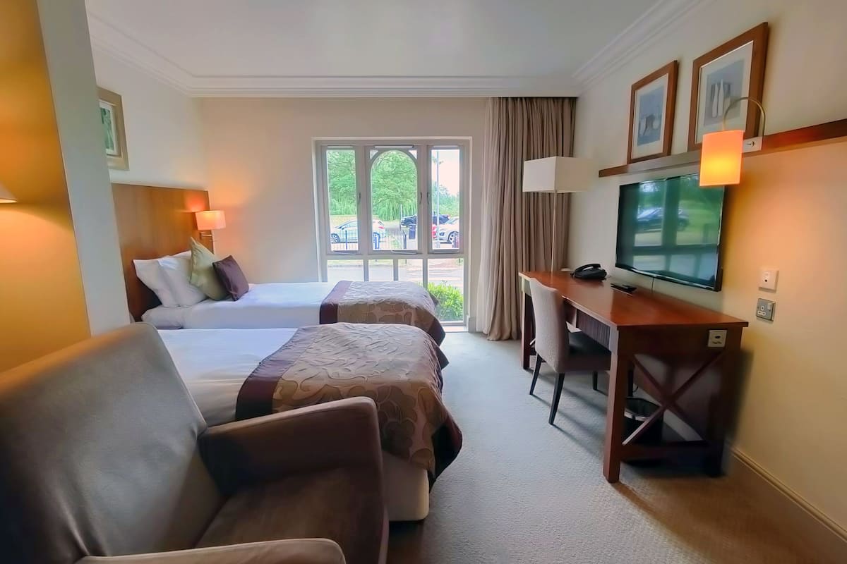 A hotel room with two beds, a desk, a television, and a view of the outdoors.
