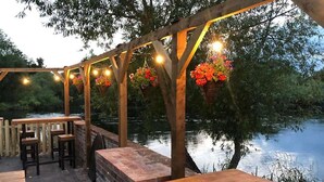 An outdoor patio with wooden tables and benches, a brick wall, hanging flower baskets, and string lights. - The Priest House Hotel (Derby)