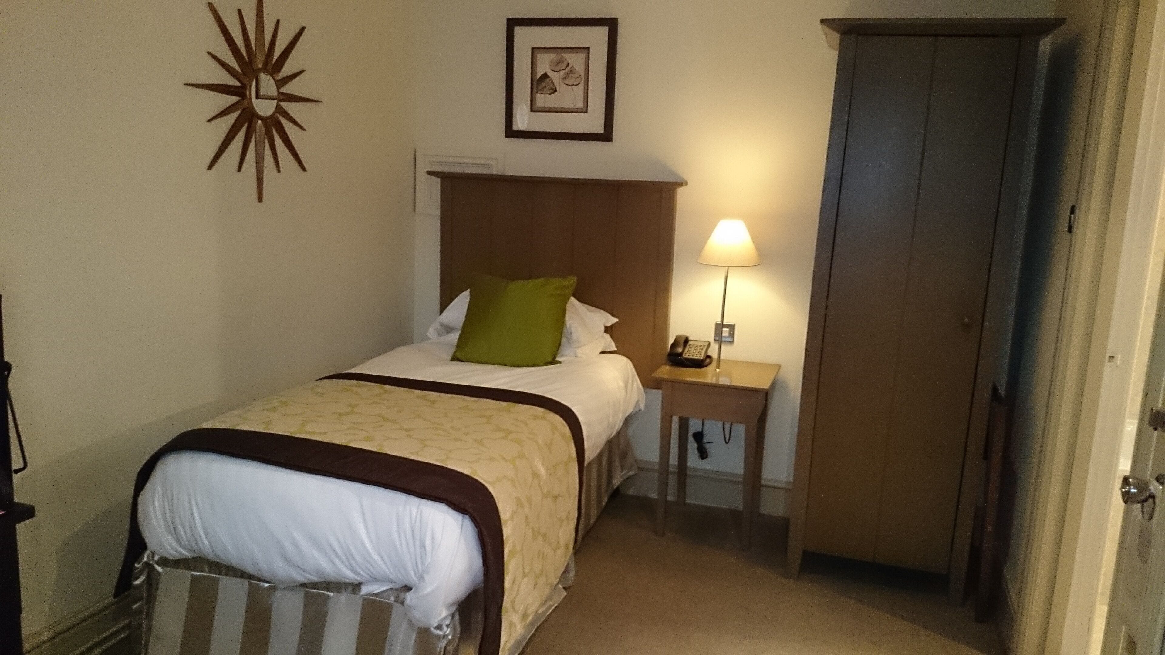 A hotel room with a bed, bedside table with a lamp, a door, and a wall clock.