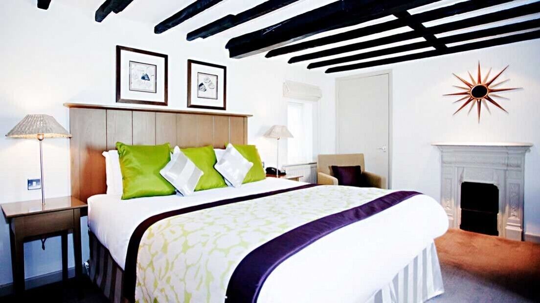 Ein Hotelzimmer mit Bett, Nachttisch, Lampe, Kamin und zwei gerahmten Bildern an der Wand.