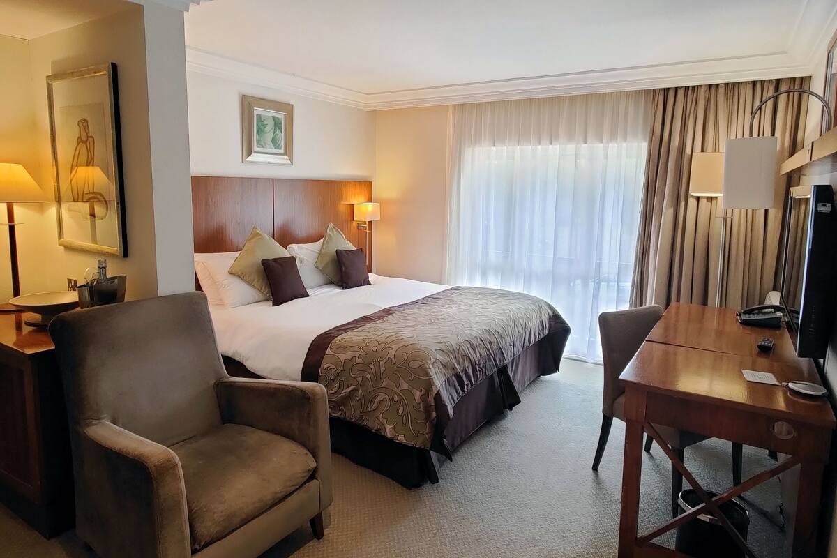 A hotel room with a large bed, two bedside lamps, a desk with a chair, a television, and a window with curtains.