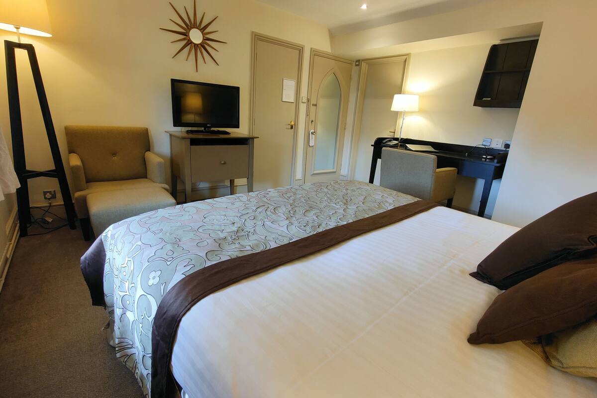 A hotel room with a bed, a chair, a desk with a computer, a TV, and a lamp.