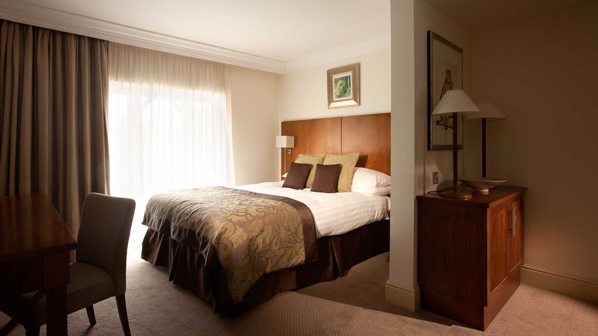 A hotel room with a bed, a desk, a chair, a lamp, and a picture on the wall.
