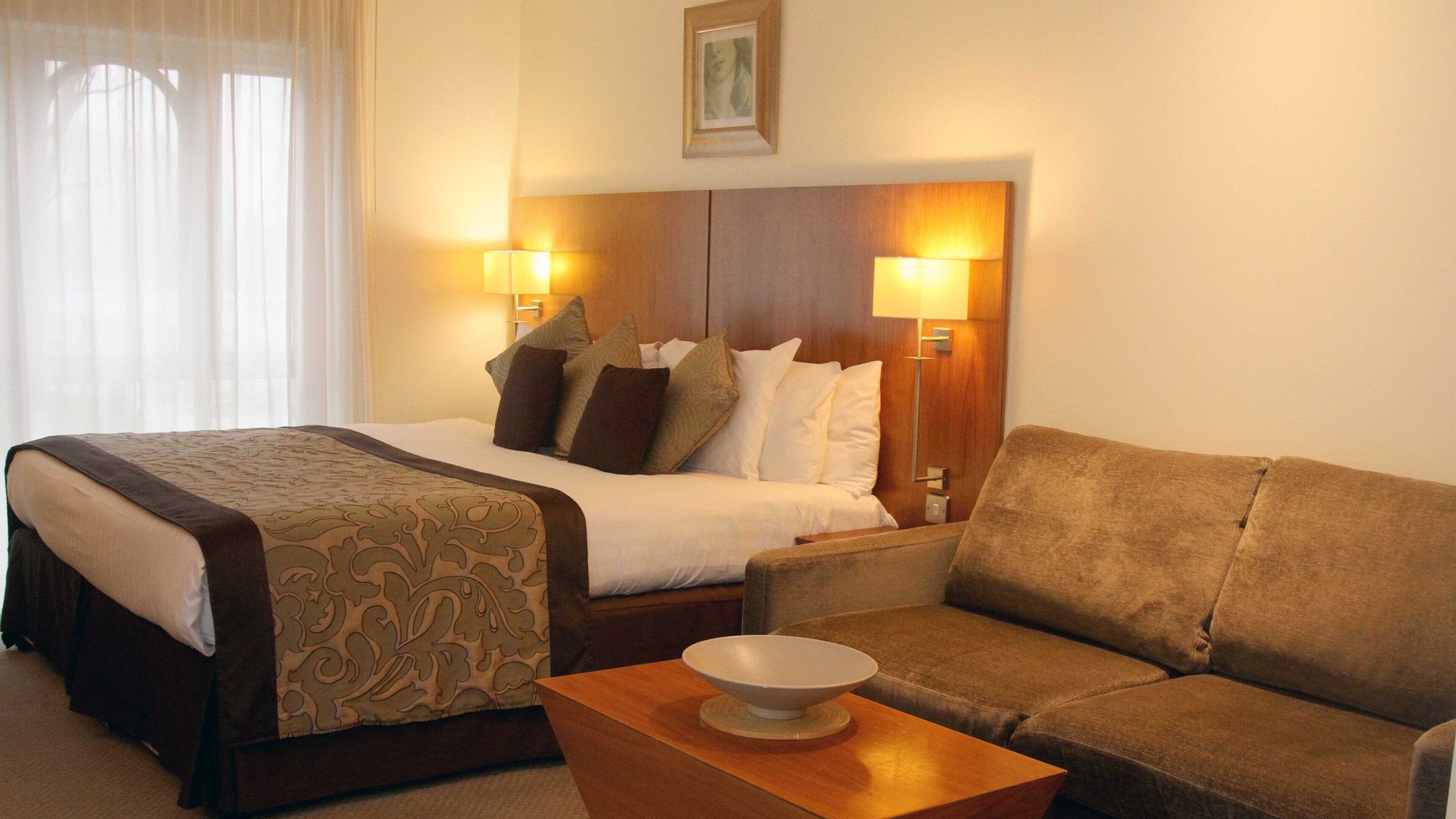 Una habitación de hotel con una cama, un sofá, una mesa con un tazón, dos lámparas de pared y un cuadro en la pared.