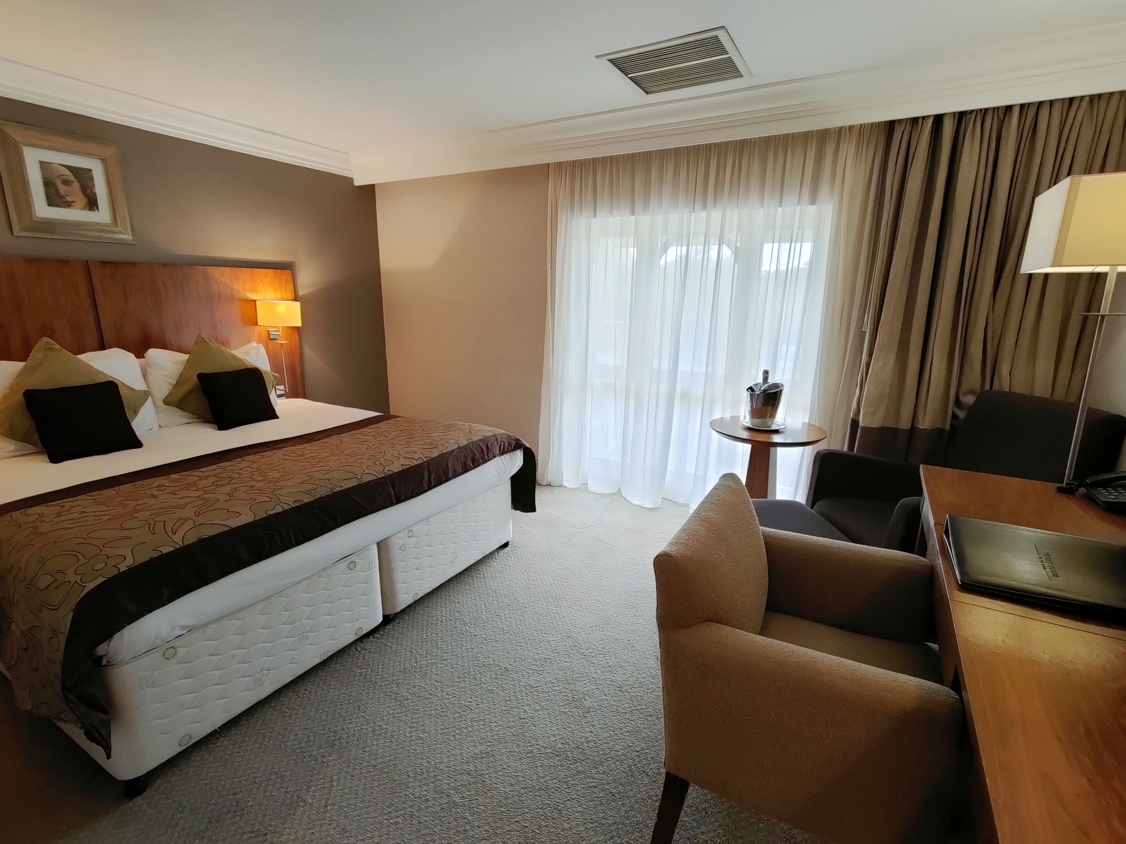 A hotel room with a bed, a desk, a chair, a lamp, a small table, and a window with curtains.