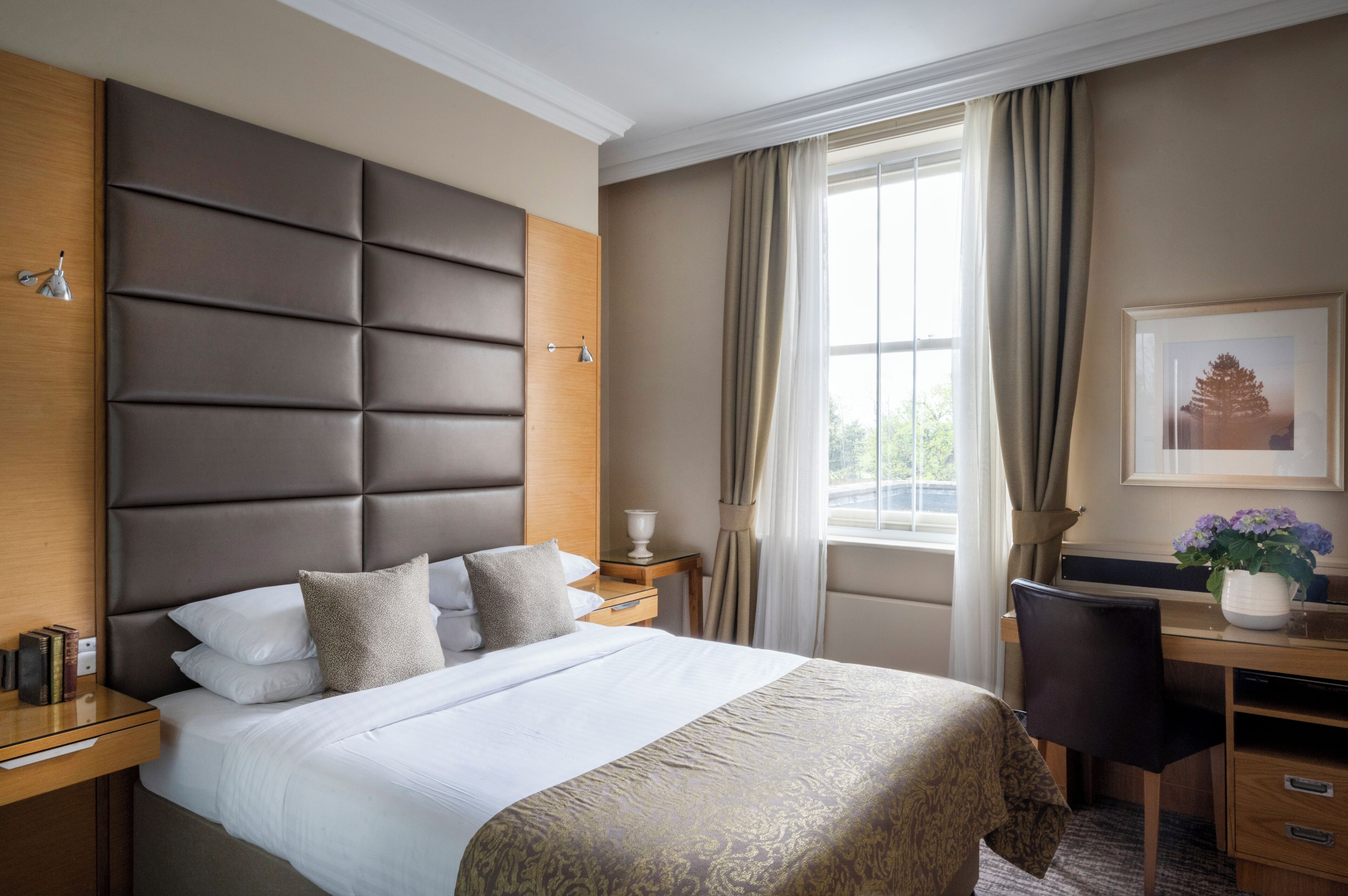 A hotel room with a large bed, a desk, a chair, a window with curtains, and a framed picture on the wall.