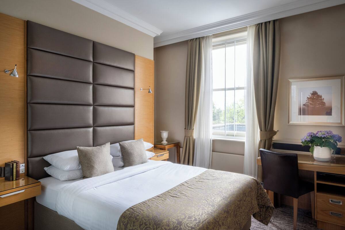 A hotel room with a large bed, a desk, a chair, a window with curtains, and a framed picture on the wall.